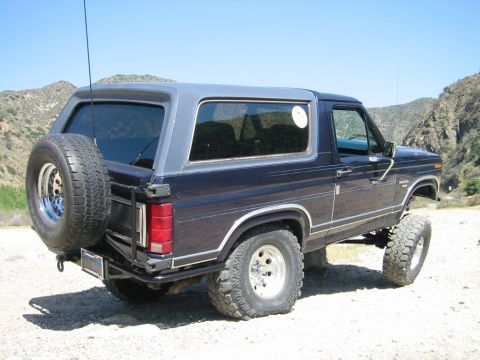 1985 Ford Bronco - This lifted Ford Bronco is parked outdoors on a dirt road with hills in the background.