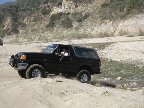 1985 Ford Bronco - A black Ford Bronco navigates a sandy terrain near a stream, with a mountainous backdrop.
