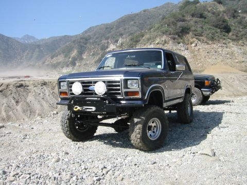1985 Ford Bronco - Two lifted Ford Broncos are parked on a gravelly surface in a mountainous, dusty area.