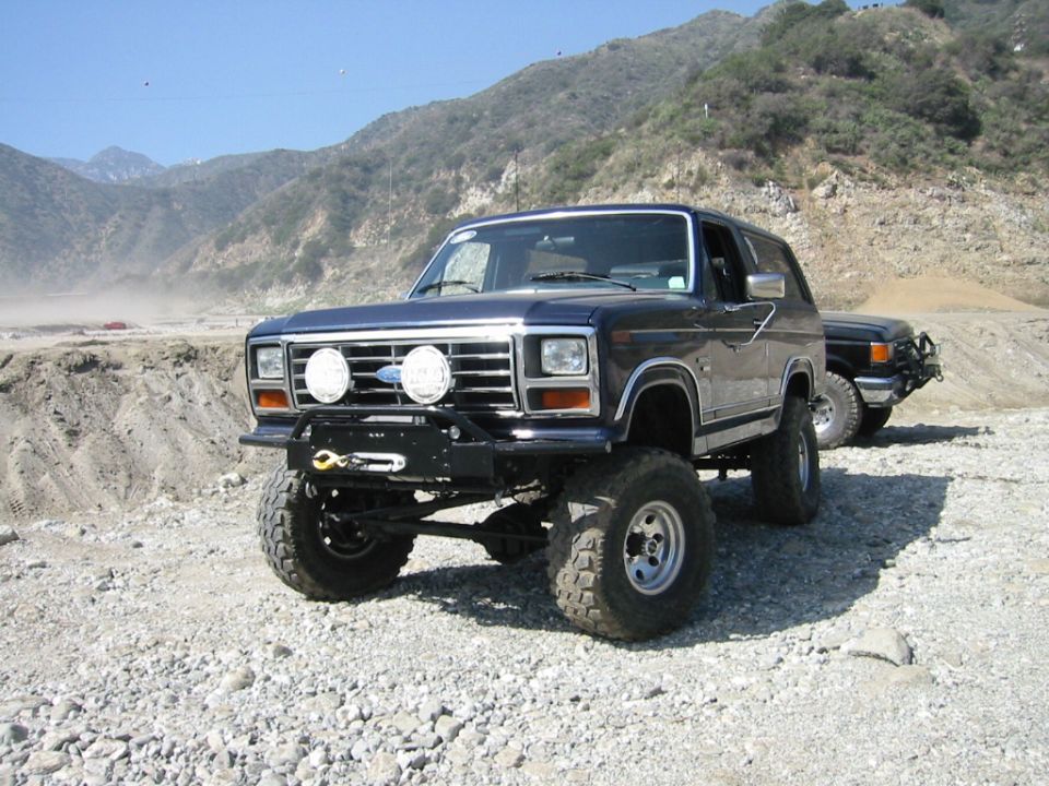 Dark lifted Ford Bronco on big off-road tires with winch bumper, parked on gravel in mountains.