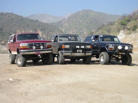 1985 Ford Bronco - Three modified Ford Broncos are lined up off-road, their lifted suspensions and tires against a mountainous backdrop.