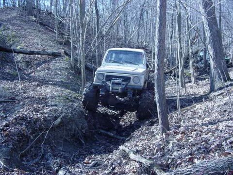 1992 Ford Bronco - A white Suzuki Samurai navigates a challenging muddy incline in a wooded area.