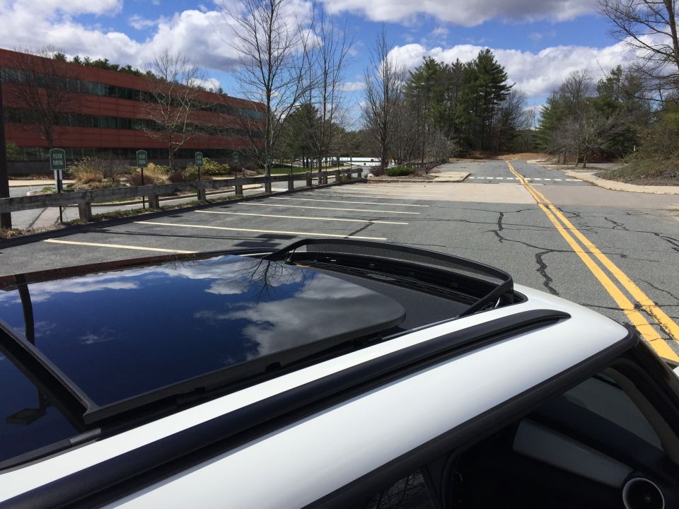 White vehicle sunroof open, reflecting clouds, with a modern building in the background.