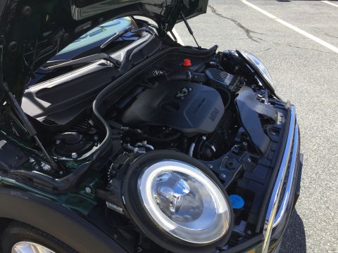 2001 Ford Crown Victoria - The open hood of a dark green Mini Cooper reveals its engine bay. The vehicle is parked on an asphalt surface with white lane markings.