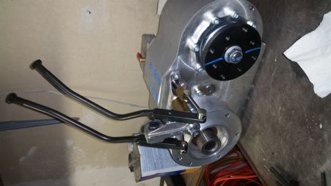 1968 Ford Bronco - A close-up view of a silver transfer case with two black shifter levers extending from the left side. The transfer case has a black circula…