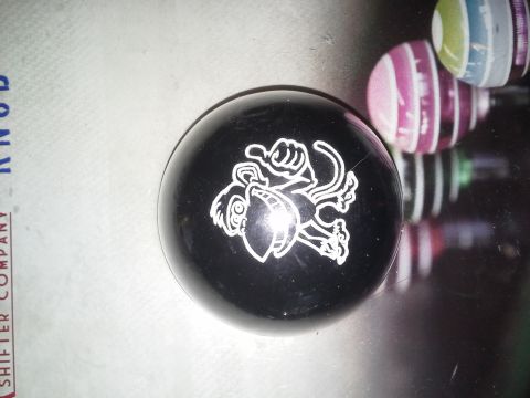 1968 Ford Bronco - A black shift knob with a white cartoon monkey design is shown on a reflective surface. The background shows reflections of colorful stripe…