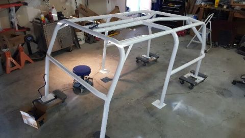 1968 Ford Bronco - A white roll cage assembly is shown in a garage setting. The structure is elevated on dollies and stands, with various tools and equipment…