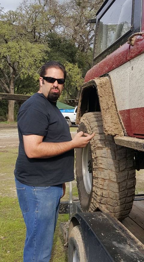 1968 Ford Bronco - A man in a black t-shirt and blue jeans stands next to a mud-covered off-road vehicle, pointing at its large tire. The vehicle appears to b…