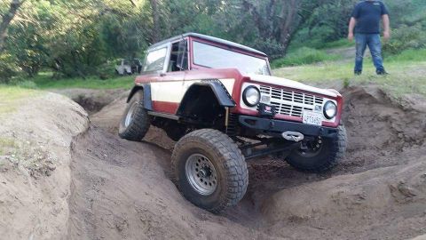 1968 Ford Bronco - A red and white Ford Bronco is navigating a steep dirt incline with a deep trench to its left. The vehicle is angled, with its front tires…