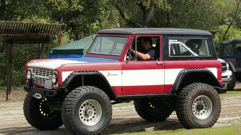 1968 Ford Bronco - A red, white, and blue Ford Bronco with large off-road tires is parked outdoors with trees and tents in the background. A person is visible…