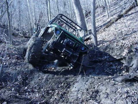 1992 Ford Bronco - An off-road vehicle navigates a steep, muddy trail through a wooded area.