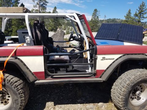1968 Ford Bronco - Side view of a red and white Ford Bronco with a roll cage, racing seats, and solar panels mounted on the hood. The vehicle is parked on a g…