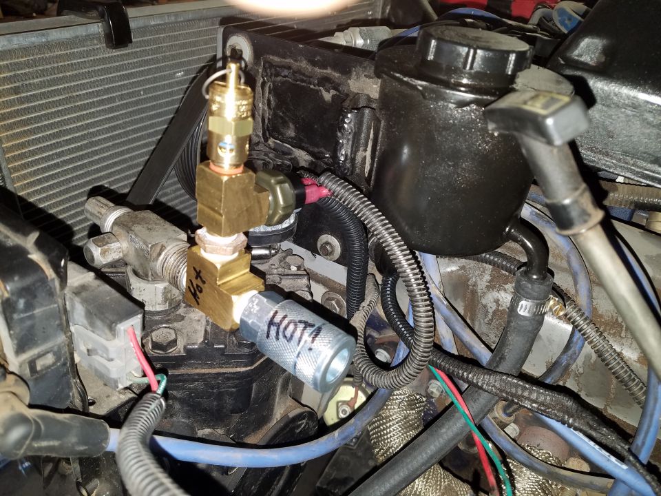 Engine bay with brass fitting labeled "HOT!", power steering reservoir, hoses, and wires.