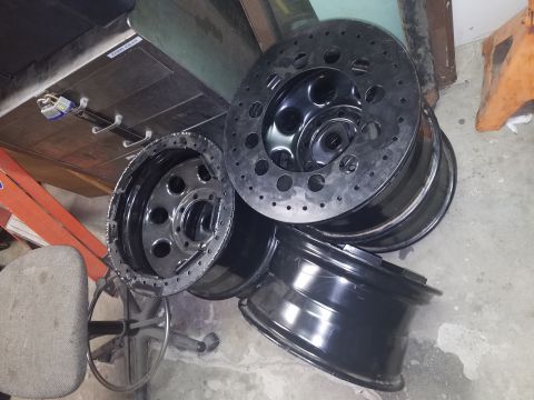 1968 Ford Bronco - A stack of four black aftermarket wheels are piled on a concrete floor. One wheel is a polished metal rim with a black center, and the othe…