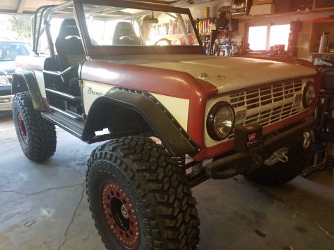 1968 Ford Bronco - A custom 1968 Ford Bronco is shown from a low front 3/4 angle in a garage. The vehicle features large off-road tires, a roll cage, and red…
