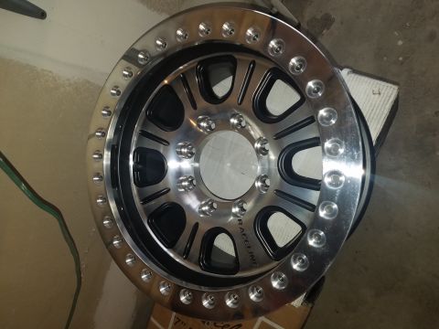 1968 Ford Bronco - A polished aluminum and black Raceline wheel with a simulated beadlock ring is shown in a close-up, angled view. The wheel is resting on a…