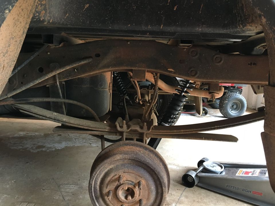 Underside of a pickup truck showing leaf springs, shock absorber, brake drum, and a jack.