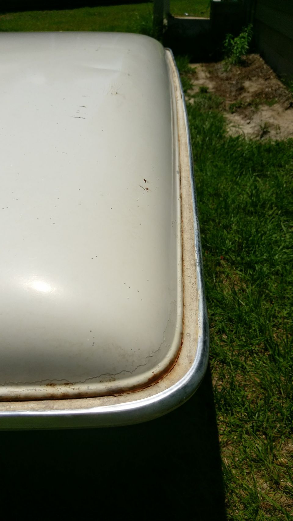 Close-up of a white vehicle's rounded corner with rusted metal trim, outdoors on grass.