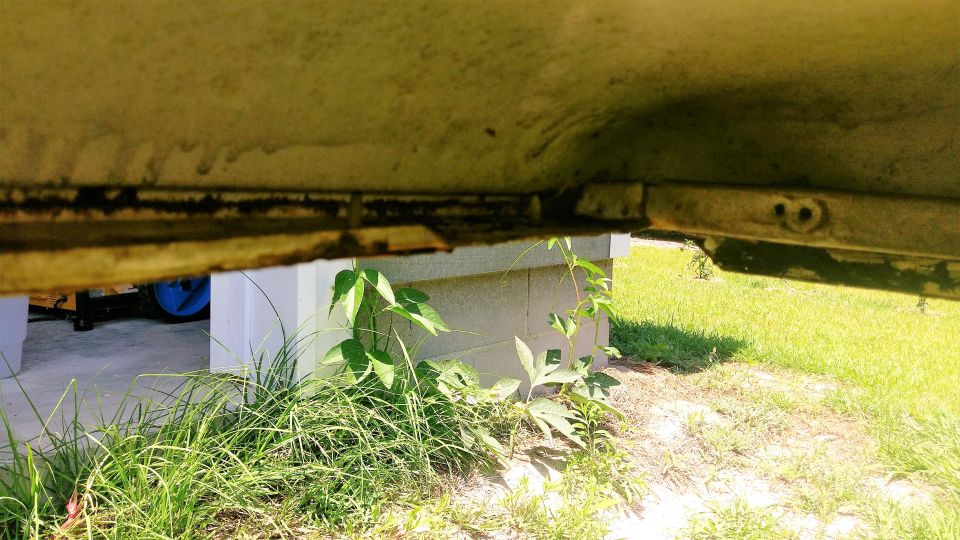 Underside of a vehicle showing rust and undercarriage components, with vegetation below.