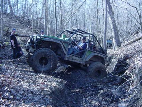 1992 Ford Bronco - An off-road vehicle navigates a steep, muddy incline in a wooded area, with another vehicle in the distance.