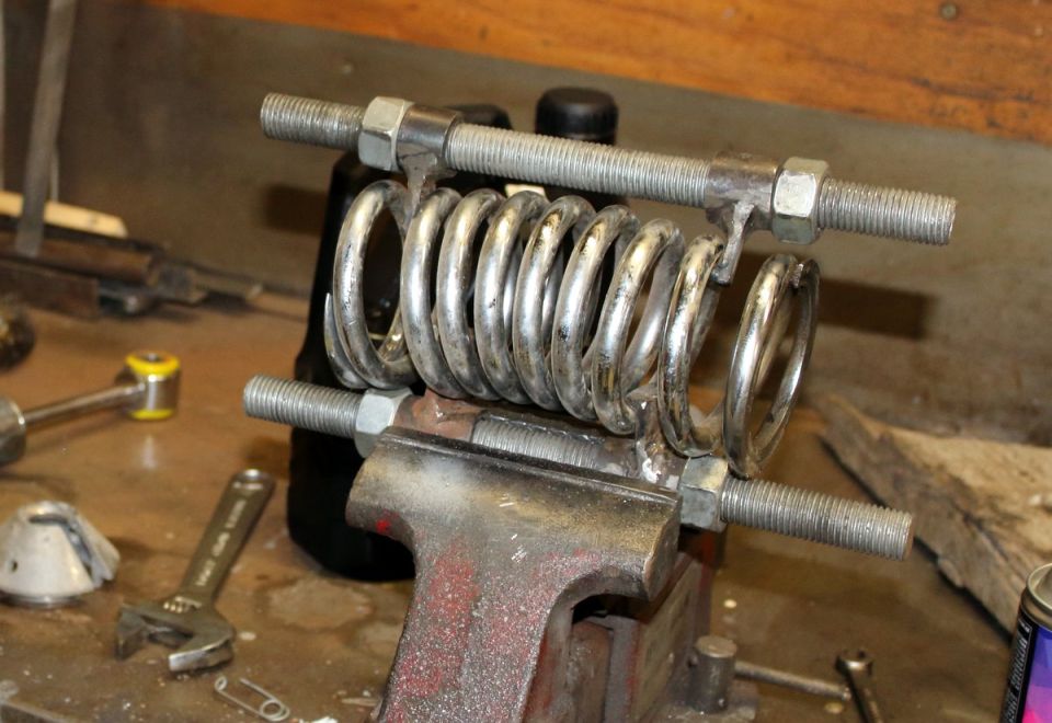 Metal spring compressed by threaded rods and nuts in a vise.