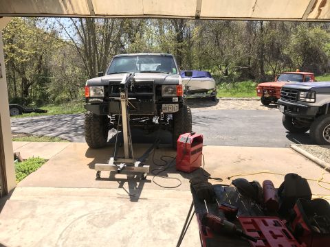 1989 Ford Bronco - A lifted, dark-colored SUV is parked in a garage or driveway with a boat on a trailer and two other trucks visible in the background. A red…