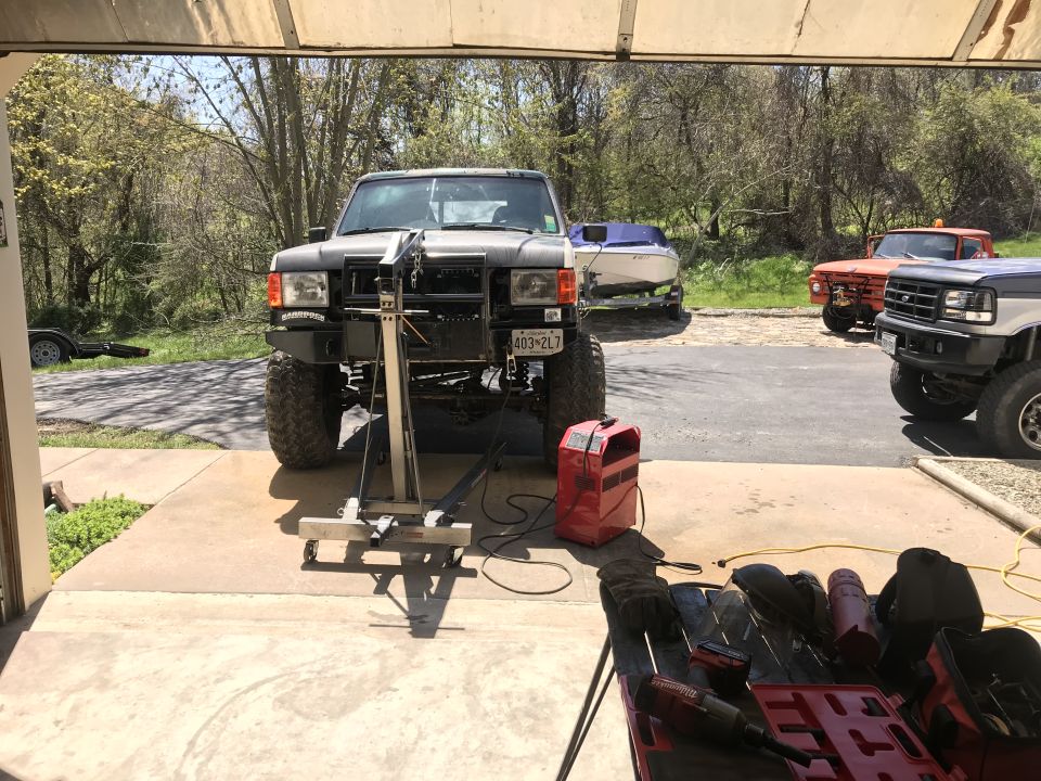 Lifted dark SUV in driveway with welding equipment and other vehicles in background.