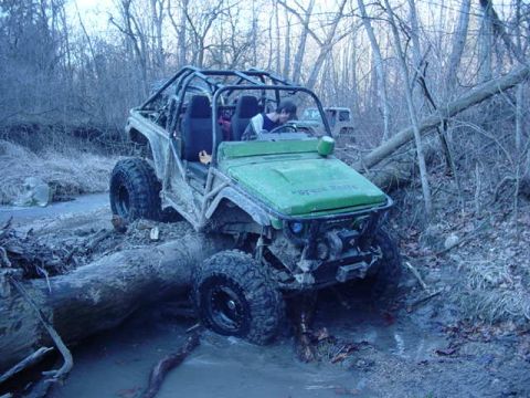 1992 Ford Bronco - A green off-road vehicle attempts to cross a muddy stream, straddling a large fallen tree.