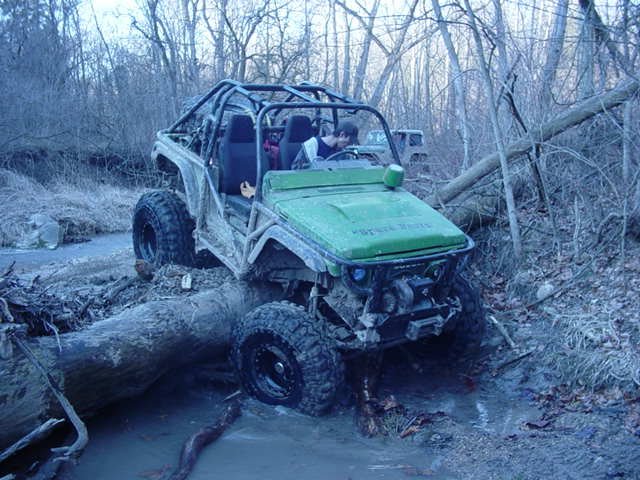Green off-road vehicle with roll cage navigating a muddy stream over a fallen log