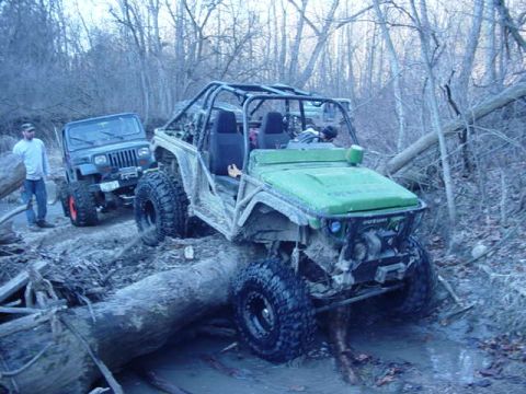 1992 Ford Bronco - Two off-road vehicles traverse a challenging muddy path, with one vehicle positioned atop a substantial fallen tree.