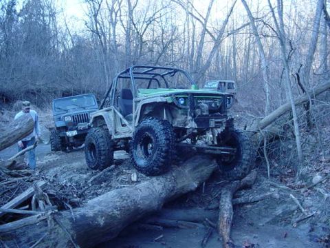1992 Ford Bronco - Two heavily modified off-road vehicles navigate a challenging muddy terrain with fallen trees.