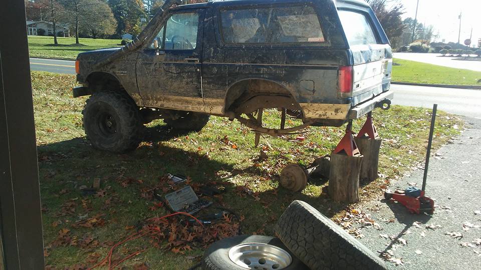 Muddy SUV on jack stands with rear axle removed, tools and spare tire nearby.