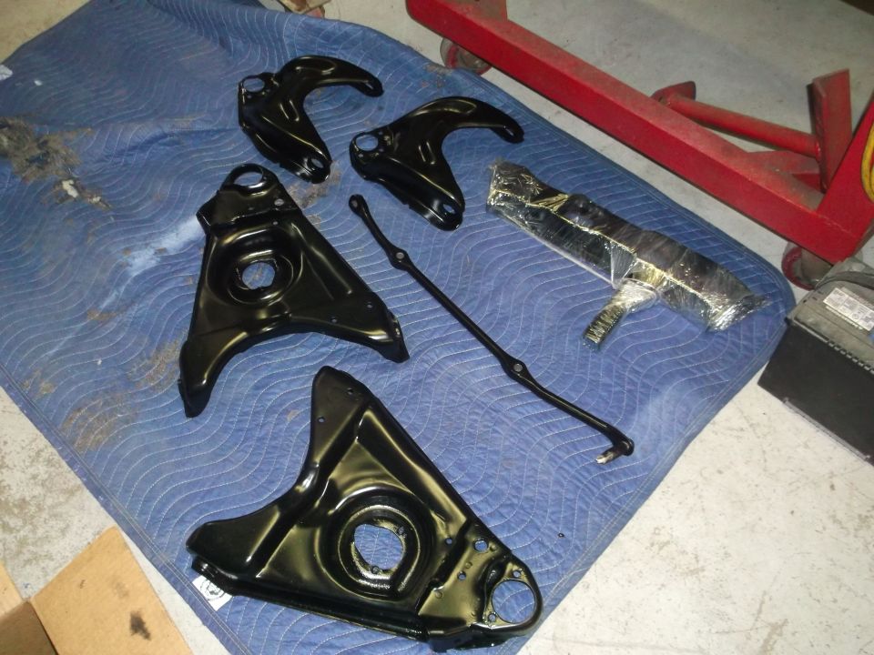 Black automotive suspension control arms and tie rod on a blue mat with a red jack stand.