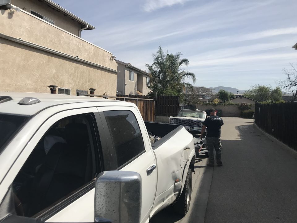 White pickup truck in foreground, white classic car on trailer behind it. Man stands near car