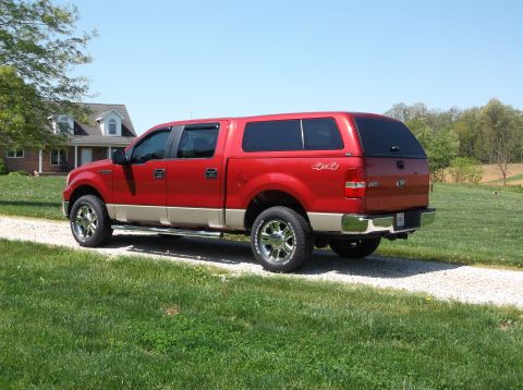 2008 Ford F-150 - A red pickup truck with a camper shell is parked on a gravel driveway in front of a house.