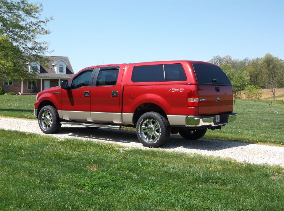 Red pickup truck with tan lower body, black camper shell, and chrome wheels parked on a gravel driveway