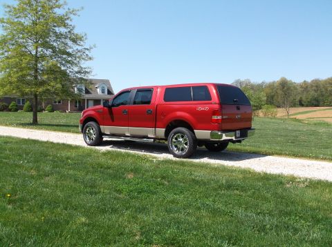 2008 Ford F-150 - A red 2008 Ford F-150 pickup truck with a tan lower body and a camper shell is parked on a gravel driveway in front of a house.