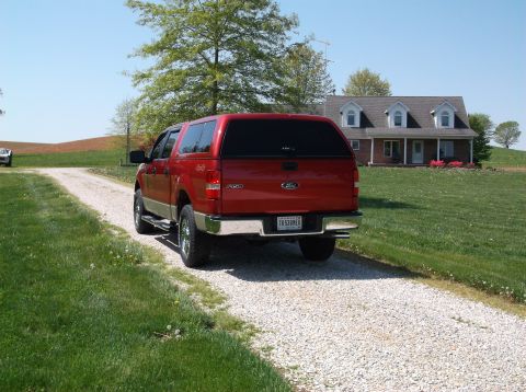 2008 Ford F-150 - A red 2008 Ford F-150 pickup truck with a camper shell is parked on a gravel driveway in front of a house.