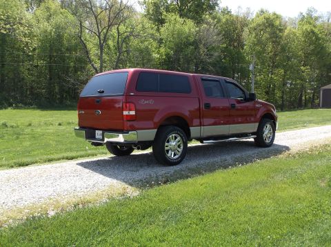 2008 Ford F-150 - A red pickup truck with a camper shell is parked on a gravel driveway, surrounded by green grass and trees.