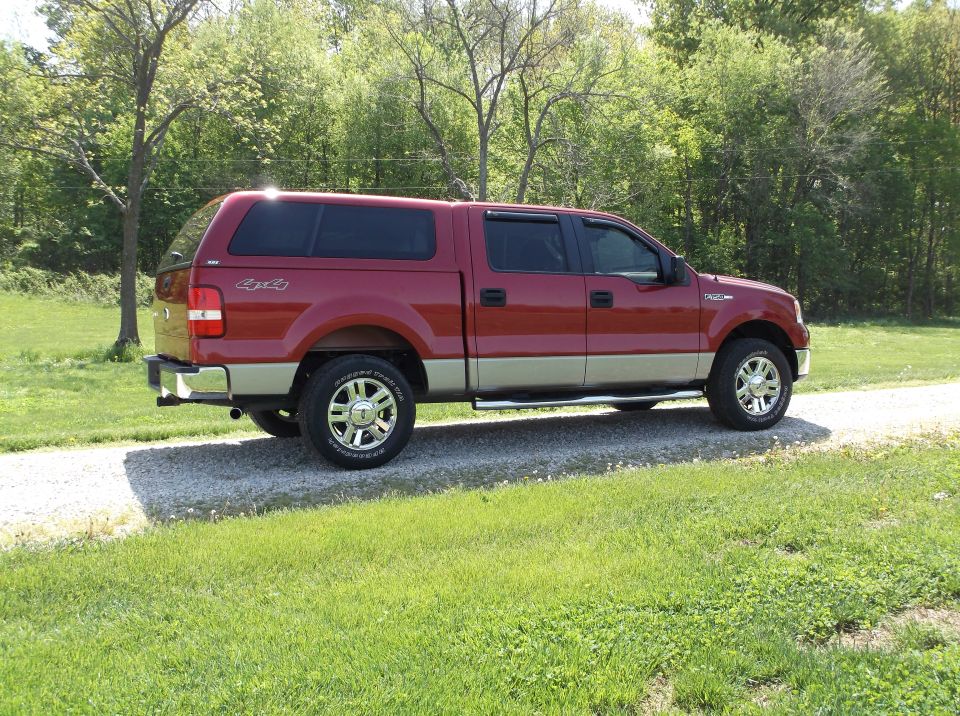 Red pickup truck with tan lower body, camper shell, and chrome wheels parked on a gravel road