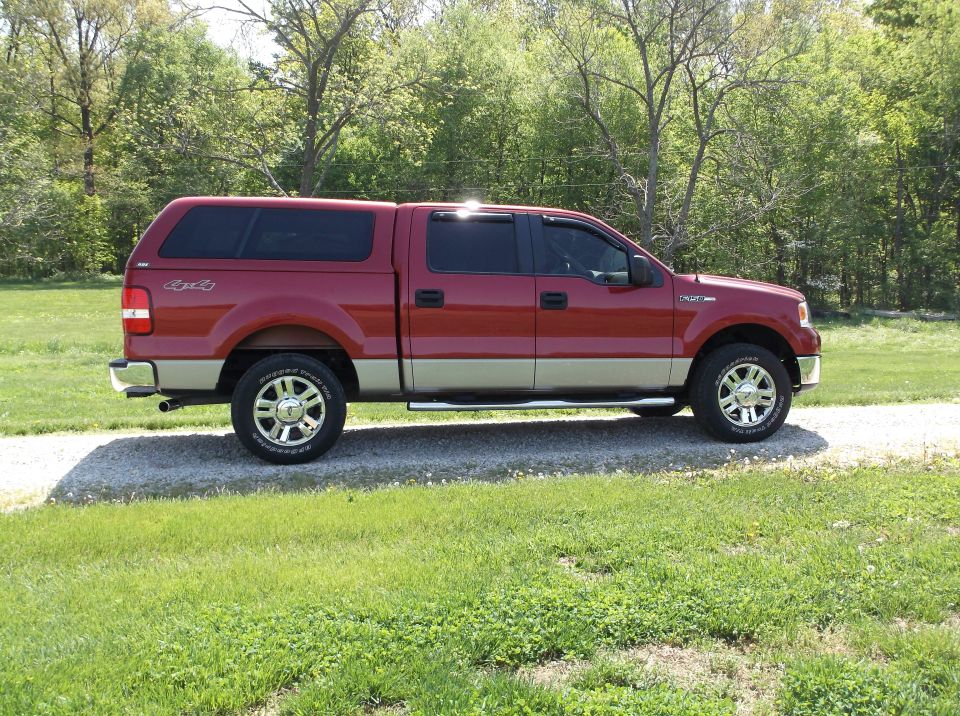 Side view of a red and tan Ford F-150 pickup truck with a camper shell parked on a gravel driveway