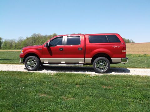 2008 Ford F-150 - A red pickup truck with a camper shell is parked on a gravel road next to a grassy field.