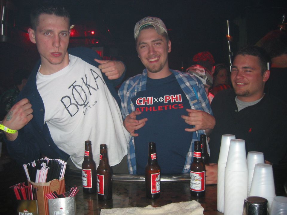Three young men posing at a bar behind a counter lined with beer bottles