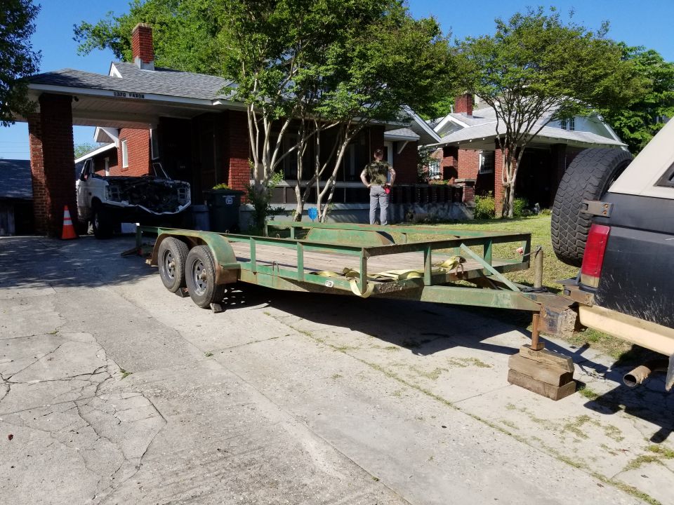 Green car trailer hitched to dark truck in driveway; stripped white vehicle with exposed engine under carport behind
