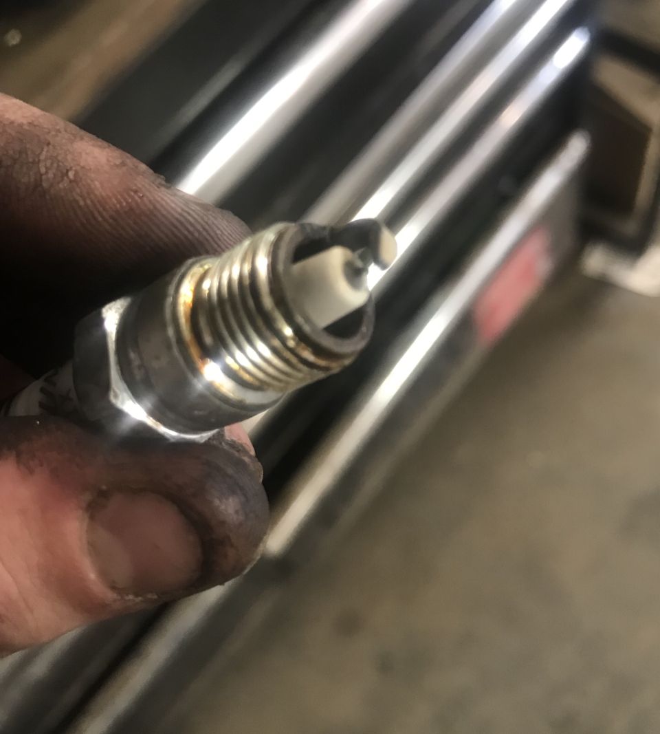 Close-up of a used spark plug being held by a dirty hand. Supermotors.net logo visible
