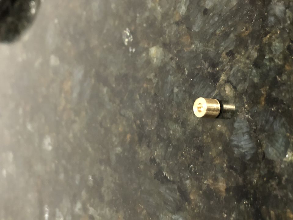 Close-up of a brass carb jet with a small pin extending from it, resting on a speckled surface. Supermotors.net logo visible