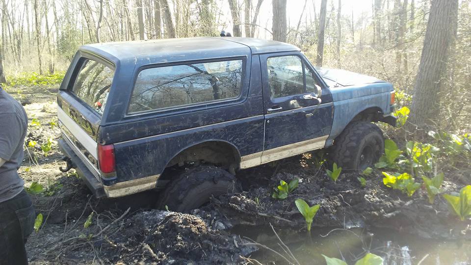 Dark blue Ford Bronco stuck in mud, right front wheel submerged, trees and foliage in background