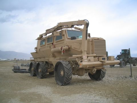 1981 Ford Bronco - A large tan armored truck with a crane is parked on a dirt lot with another vehicle in the background.