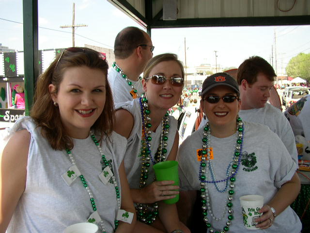 Three women wearing green beads and shamrock decorations smile for the camera at an outdoor event