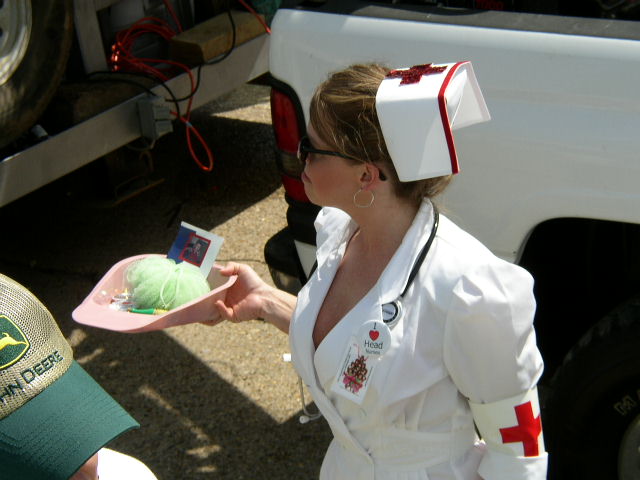 Person in nurse costume with stethoscope and red cross hat holds tray with green yarn and a small photo